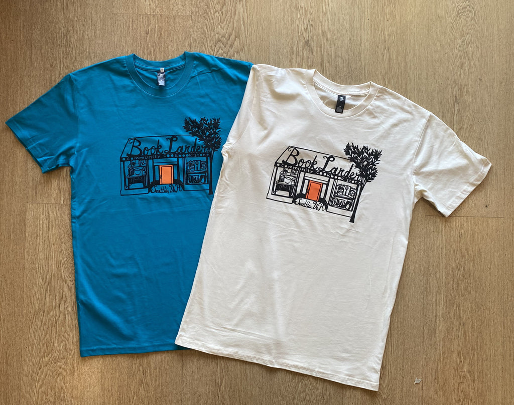 Two t-shirts, one teal and one meringue, with a graphic design on a wooden surface.